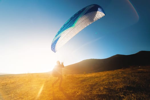 A paraglider above the ground is preparing to fly. A professional athlete raised his parachute over a yellow field against the backdrop of hills on a sunny day
