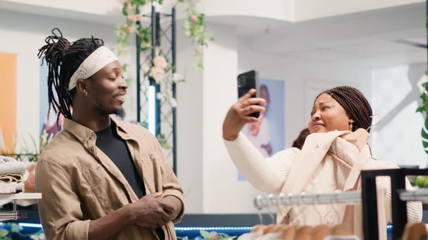 Man in fancy fashion boutique convincing his girlfriend to buy stylish formal clothes on hangers. Customers in premium showroom debating whether to purchase new designer clothing collection or not