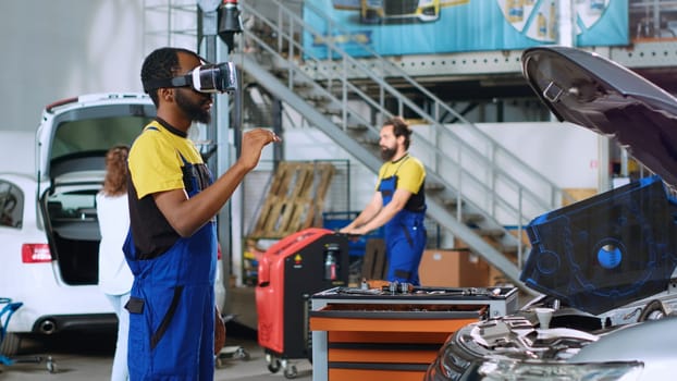 Mechanic in repair shop using advanced virtual reality technology to visualize car mechanical component in order to fix it. BIPOC garage employee wearing vr goggles while mending broken vehicle