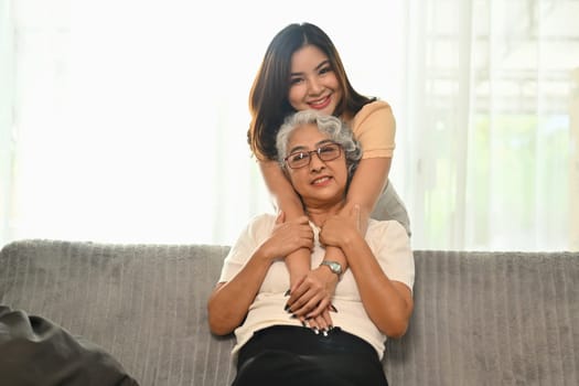 Adult daughter standing behind couch and hugging older mother. Family bonding, International hug day concept.