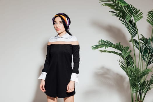 a woman in a black dress and scarf stands against a white wall and a green plant