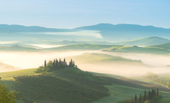 House surrounded by cypress trees among the misty morning sun-drenched hills of the Val d'Orcia valley at sunrise in San Quirico d'Orcia, Tuscany, Italy