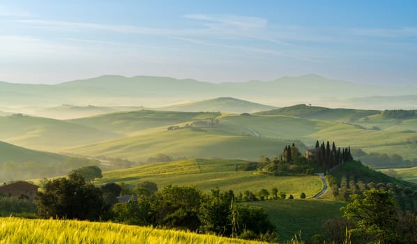 House surrounded by cypress trees among the misty morning sun-drenched hills of the Val d'Orcia valley at sunrise in San Quirico d'Orcia, Tuscany, Italy