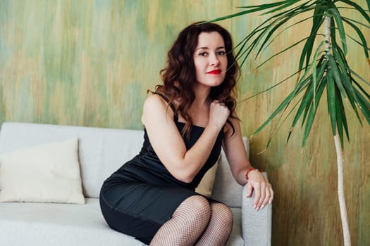 brunette woman in a black dress sits on the couch by a green plant in a room