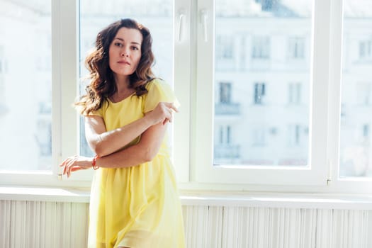 beautiful brunette woman in yellow dress stands in a bright room by the window