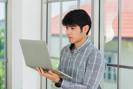 Asian businessman smiling and working on a laptop computer stand near window in office. Portrait of handsome man standing and online meeting with computer notebook in office lobby