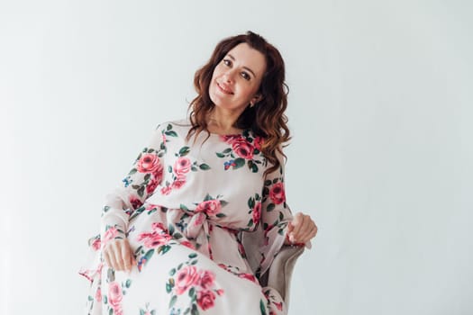 brunette woman in a floral dress sits in a chair on a light background