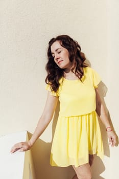 brunette woman in a yellow dress stands against a beige wall