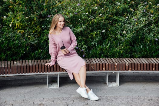 blonde woman sitting on bench with a cup of drink by the green bushes