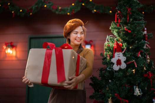 Happy woman wearing Santa hat holding of gift box. Positive emotional Santa girl. with a beautifully decorated Christmas tree serving as the background. festive Xmas concept.