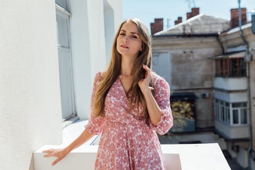 a blonde in a pink dress in a flower stands on the balcony in the apartment house