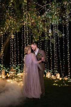 the first wedding dance of the bride and groom in the glade of the country club in the light of sunset and warm garlands