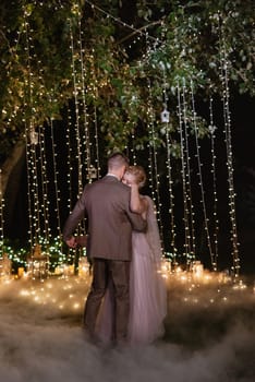 the first wedding dance of the bride and groom in the glade of the country club in the light of sunset and warm garlands