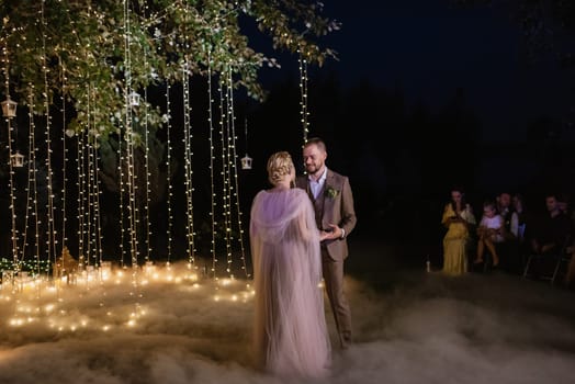 the first wedding dance of the bride and groom in the glade of the country club in the light of sunset and warm garlands