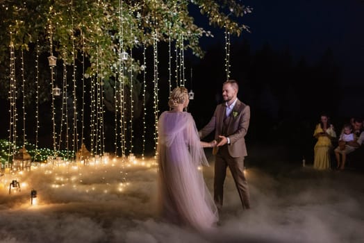 the first wedding dance of the bride and groom in the glade of the country club in the light of sunset and warm garlands