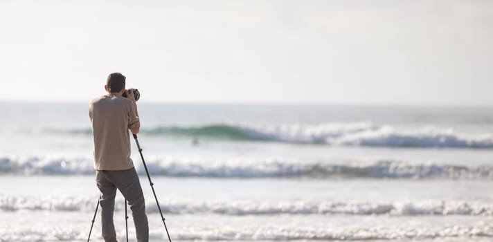 Young handsome photographer with a tripod and a camera on the beach - view from behind