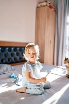 Little girl with a comb in her hands sits next to the cat on the bed. High quality photo