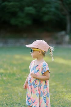 Little girl in sunglasses stands on the lawn looking into the distance. Side view. High quality photo