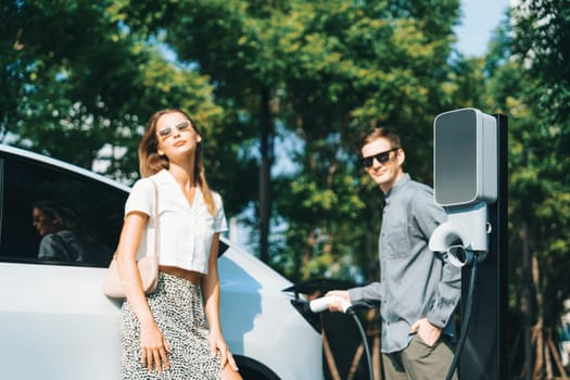 Young couple travel with EV electric car charging in green sustainable city outdoor garden in summer shows urban sustainability lifestyle by green clean rechargeable energy of electric vehicle innards