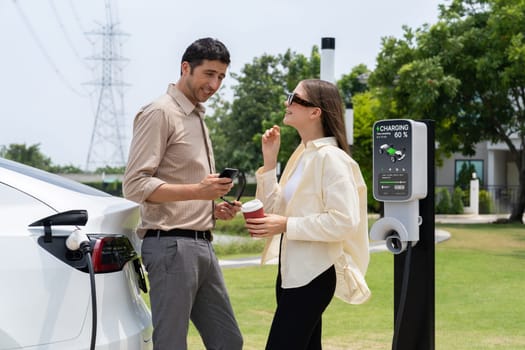 Young couple with coffee pay for electricity while recharge EV car battery at charging station connected to electrical power grid tower as electric industrial for eco friendly car travel. Expedient