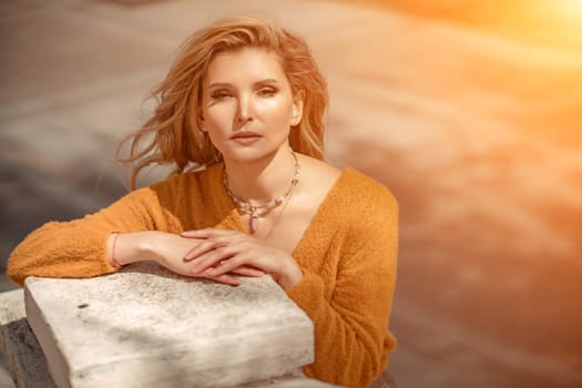 A middle-aged woman looks like a good blonde with curly beautiful hair and makeup on the background of the building. She is wearing a yellow sweater