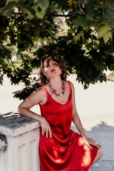 woman in a red silk dress and a bandage on her head smiles against the background of the leaves of a tree. She is leaning on the coop and looking into the camera. Vertical photo