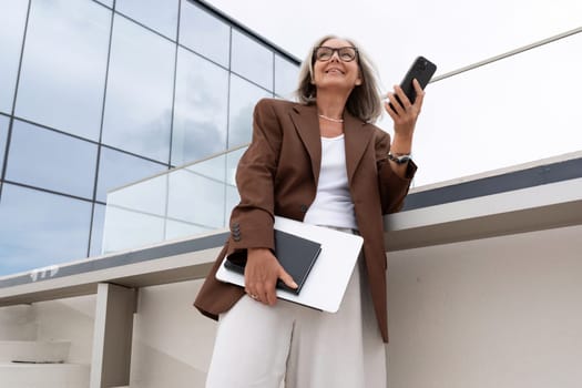 a gray-haired mature business lady dressed in a stylish jacket and trousers looks successfully holding a smartphone and documents in her hands on the street