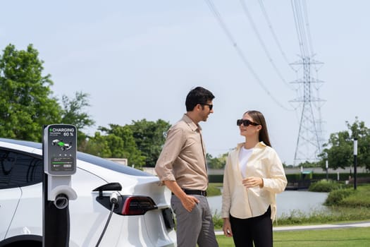 Young couple recharge EV car battery at charging station connected to power grid tower electrical industrial facility as electrical industry for eco friendly vehicle utilization. Expedient