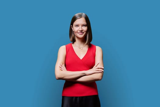 Portrait of young confident woman in red on blue studio background. Successful fashionable female with crossed arms looking at camera. Business, work, services, education, fashion beauty professions