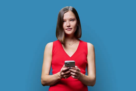 Young woman with smartphone in hands on blue studio background. Smiling successful fashionable female looking at camera. Using mobile applications for work business shopping banking study leisure