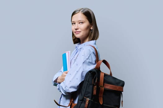 Portrait of young university student with backpack of textbooks on gray studio background. Smiling confident positive 20s female looking at camera. Education, knowledge, lifestyle, youth concept