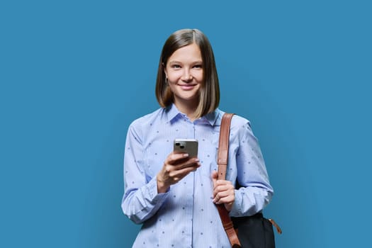 Young woman student with smartphone backpack on blue studio background. Smiling attractive female looking at camera. Using mobile applications apps for work business study leisure, technology, people