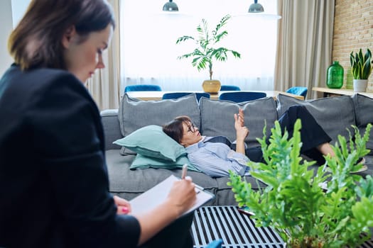 Female psychologist is having therapy session with middle-aged woman patient lying on sofa in office. Psychology, psychotherapy, therapy, counseling, treatment, mental health concept