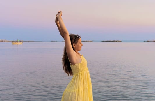 An attractive model in a yellow dress poses with her hands raised above her head, waiting for the sunrise on the sea.