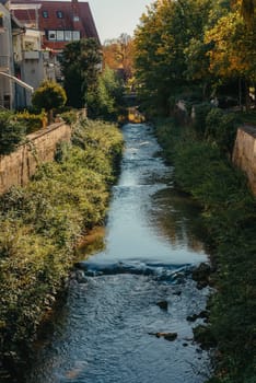 Beautiful small river with clean and clear water front of colorful autumn trees and small old town on the hill agaist nice blue and clouds sky during autumn in Europe.