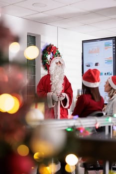 Team leader dressed as Santa Claus holding Christmas thematic meeting with employees in festive decorated office. Management executive analyzing company statistic charts during winter holiday season