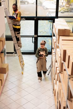 Storehouse manager analyzing clients orders checklist on tablet, scanning merchandise barcode using store scanner before preparing delivery. Supervisor in protective overall working in warehouse