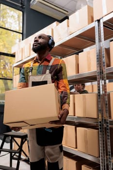 Supervisor working at merchandise distribution in storage room, carrying cardboard boxes while listening music. Storehouse manager wearing headphones, preparing customers packages for delivery