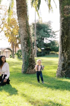 Little laughing girl with a teddy bear walks on a sunny lawn past a smiling woman. High quality photo