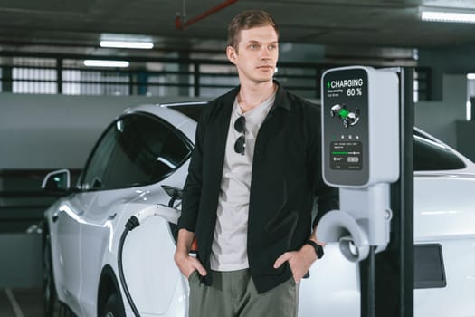 Young man travel with EV electric car to shopping center parking lot charging in downtown city showing urban sustainability lifestyle by green clean rechargeable energy of electric vehicle innards