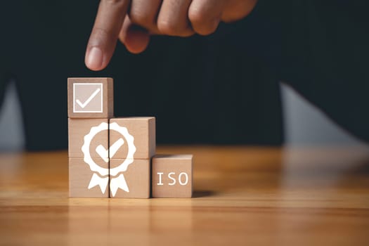 A hand holding wooden cubes with ISO icons symbolizes the concept of quality control and assurance. An ISO banner in the background represents international standards and certification.