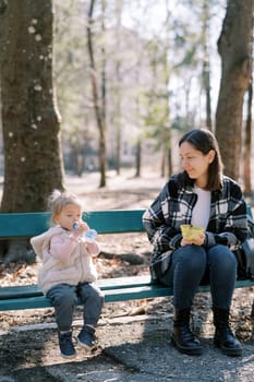 Little girl drinks water from a bottle while sitting next to her mother on a bench in a sunny park. High quality photo