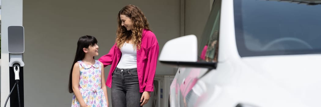 Happy little young girl learn about eco-friendly and energy sustainability as she help her mother recharge electric vehicle from home EV charging station. EV car and modern family. Panorama Synchronos