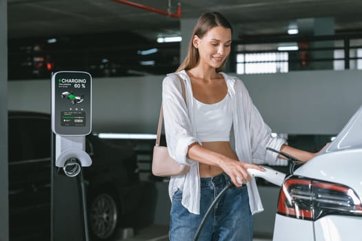 Young woman travel with EV electric car to shopping center parking lot charging in downtown city showing urban sustainability lifestyle by green clean rechargeable energy of electric vehicle innards