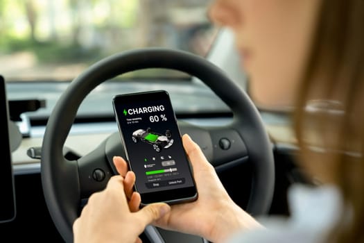 Young woman inside the vehicle, checking EV vehicle's application on battery recharging electricity status display on smartphone screen in modern EV car on her road trip journey. Exalt