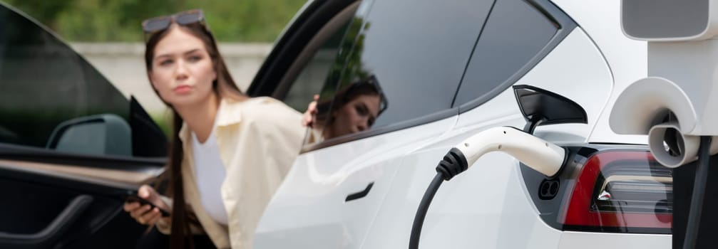 Young woman use smartphone to pay for electricity at public EV car charging station green city park. Modern environmental and sustainable urban lifestyle with EV vehicle. Panorama Expedient