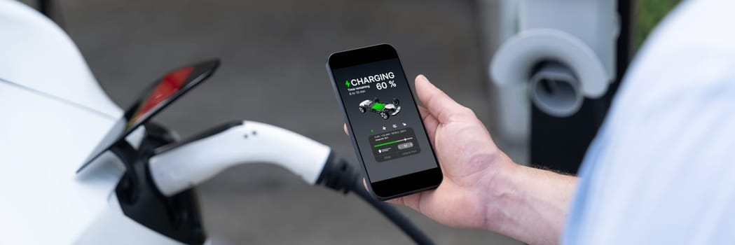 Modern eco-friendly man recharge electric vehicle from EV charging station. EV technology utilization for tracking energy usage to optimize battery charging display on smartphone. Panorama Synchronos