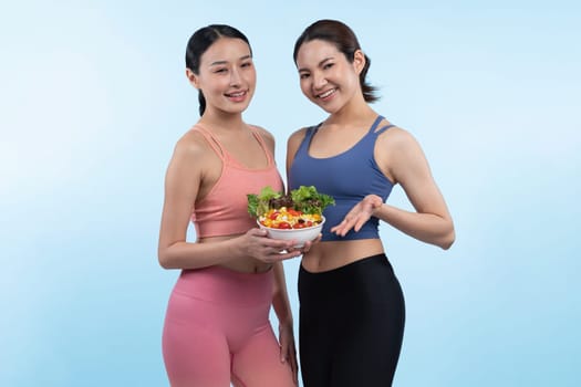 Two young sporty Asian women in sportswear holding salad bowl fill with fruit and vegetable. Natural youthful and fit body lifestyle people with balance nutrition on isolated background. Vigorous