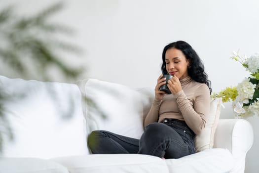 Happy woman drinking coffee on a sofa at home for crucial rest and relaxation. Portrait of young African American woman holding a cup.