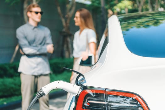 Young couple travel with EV electric car charging in green sustainable city outdoor garden in summer shows urban sustainability lifestyle by green clean rechargeable energy of electric vehicle innards